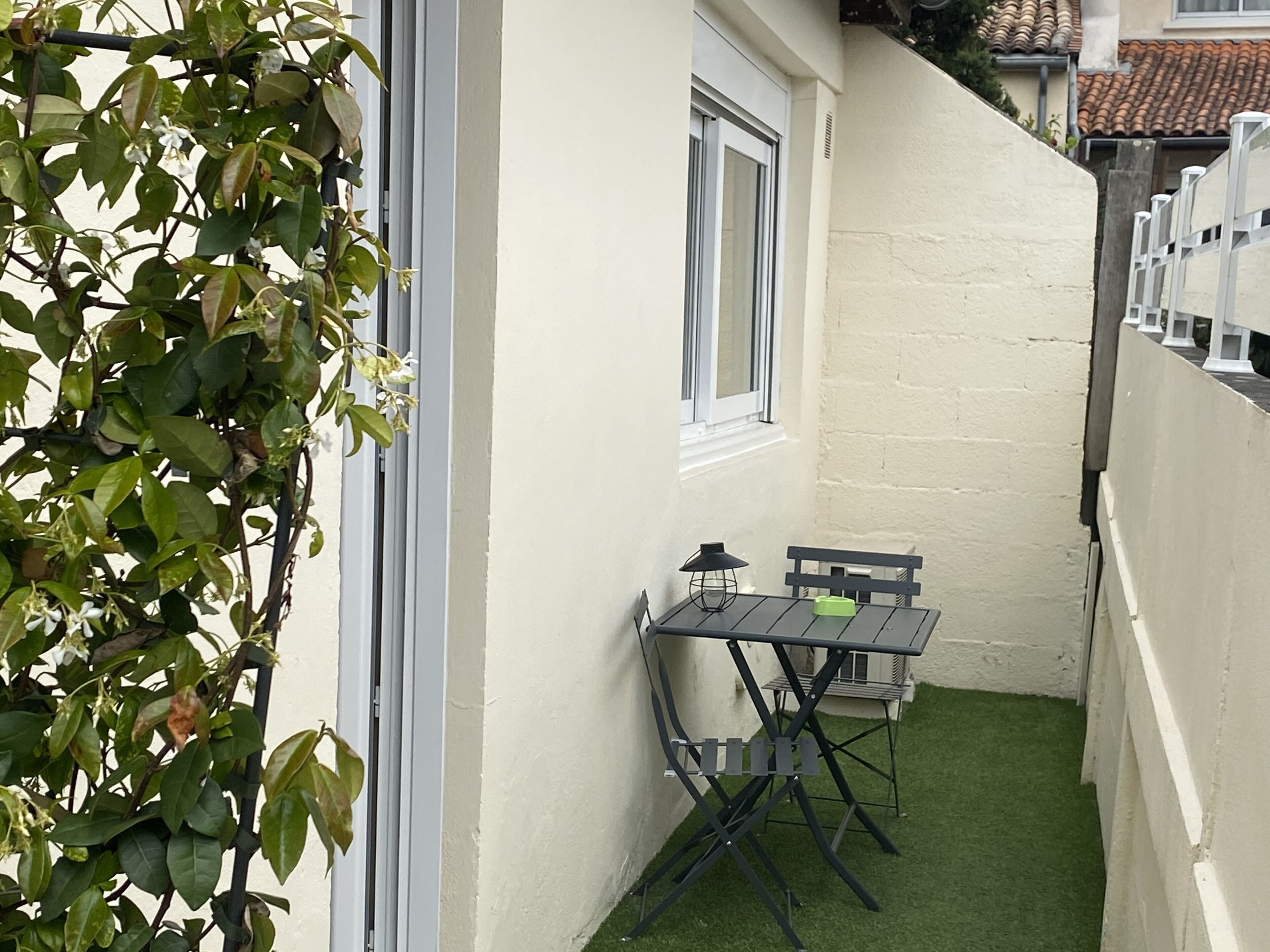 Une petite terrasse extérieure avec une table et des chaises, entourée d’un mur blanc et d’une zone recouverte de gazon synthétique vert.