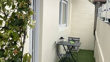Une petite terrasse extérieure avec une table et des chaises, entourée d’un mur blanc et d’une zone recouverte de gazon synthétique vert.