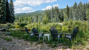 A lakeside seating area with chairs and tables, surrounded by dense forest and a mountain in the background.