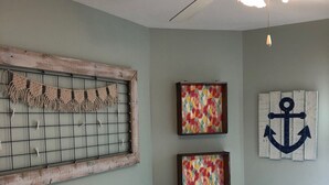A bedroom with a white bed, a ceiling fan, and wall decorations including a macrame piece, colorful paintings, and an anchor artwork. - Adorable 2BR Condo on Long Beach Island LBI  in Ship Bottom. Beach Block! (Ship Bottom)