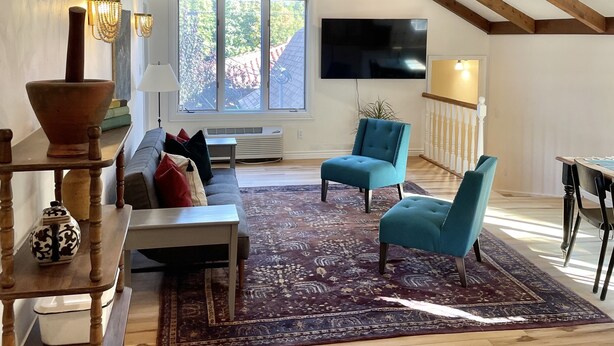 A spacious living room with a high ceiling, exposed wooden beams, a large window, a flat-screen TV, and a teal armchair.