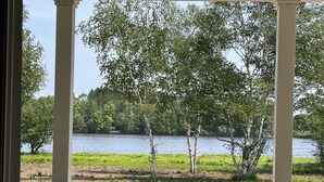A view from a porch with a white railing, overlooking a lake surrounded by trees and grassy areas.