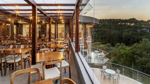 A restaurant with wooden tables and chairs, a glass railing, and a view of a forested area. - Laghetto Resort Golden (Gramado)