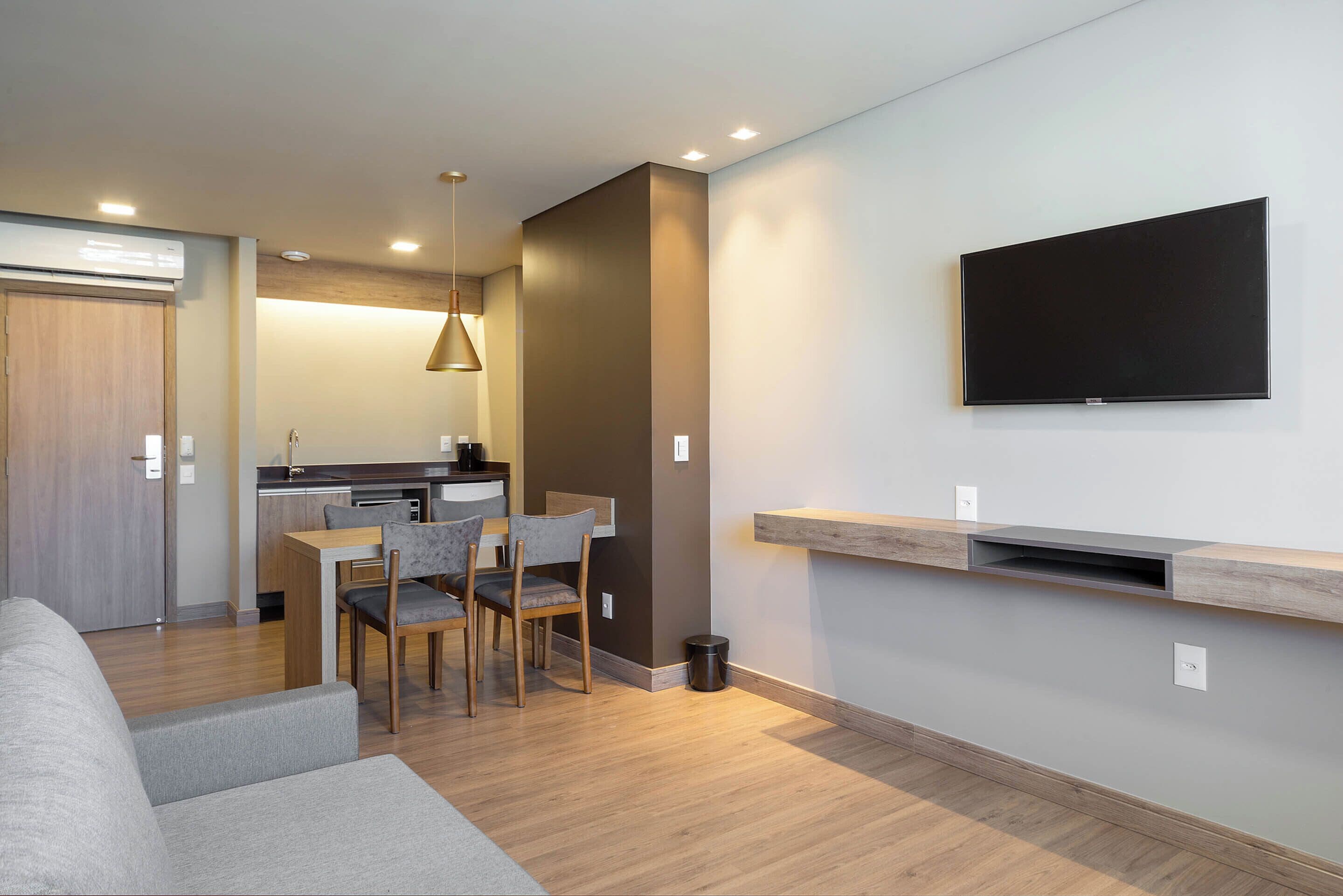 A modern living room with a grey sofa, a dining area with a table and chairs, a kitchen in the background, and a mounted TV on the wall.