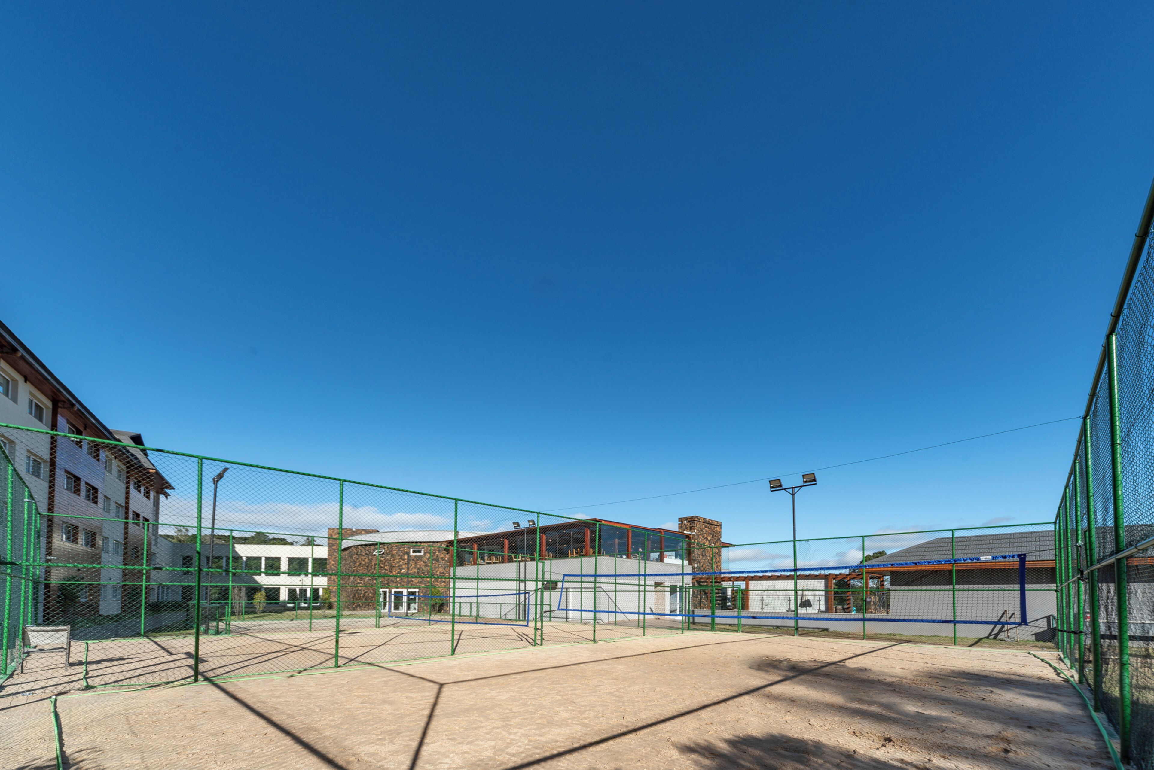 Una cancha de tenis con red, rodeada por una cerca verde, y edificios al fondo.