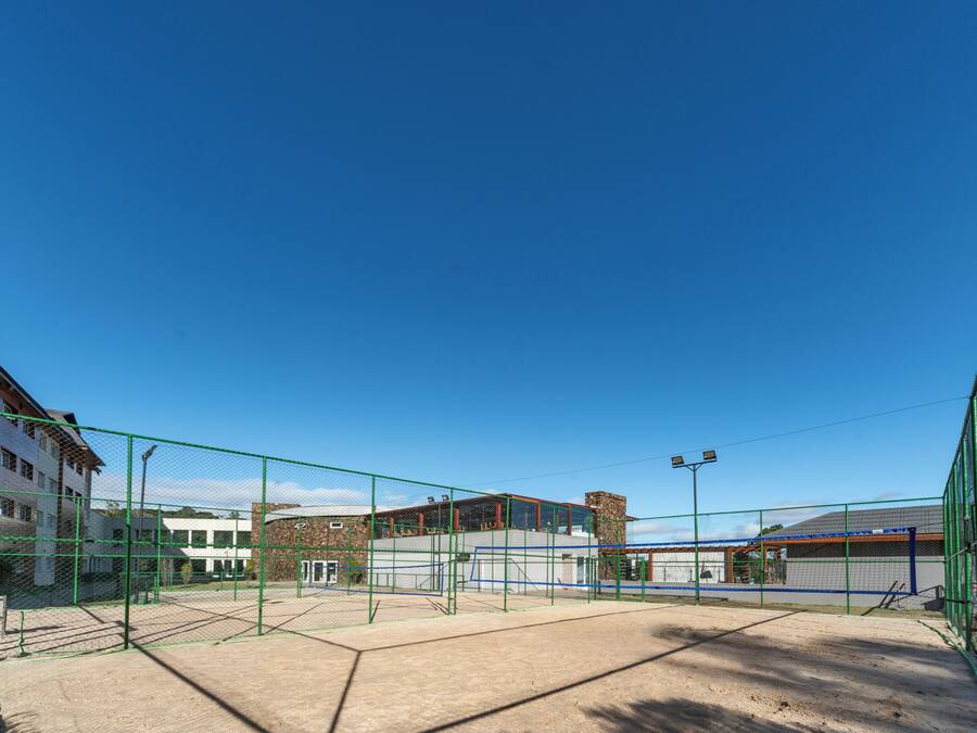 Una cancha de tenis con red, rodeada por una cerca verde, y edificios al fondo.
