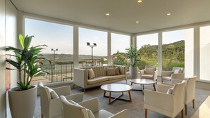 A modern living room with a beige sofa, white armchairs, and a coffee table. Large windows offer a view of a green landscape. - Laghetto Resort Golden (Gramado)