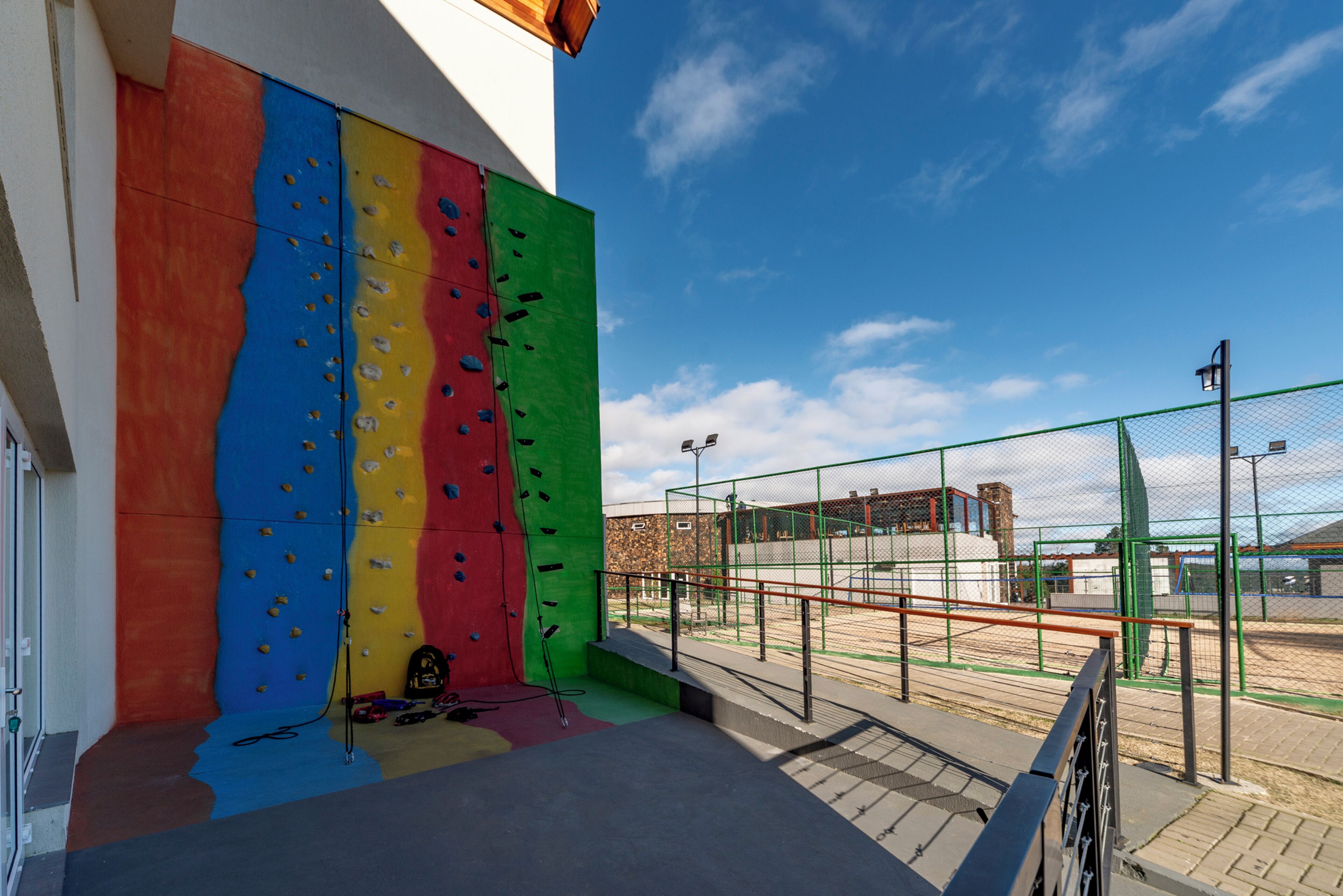 A climbing wall with colorful panels and a fenced area with a basketball court.