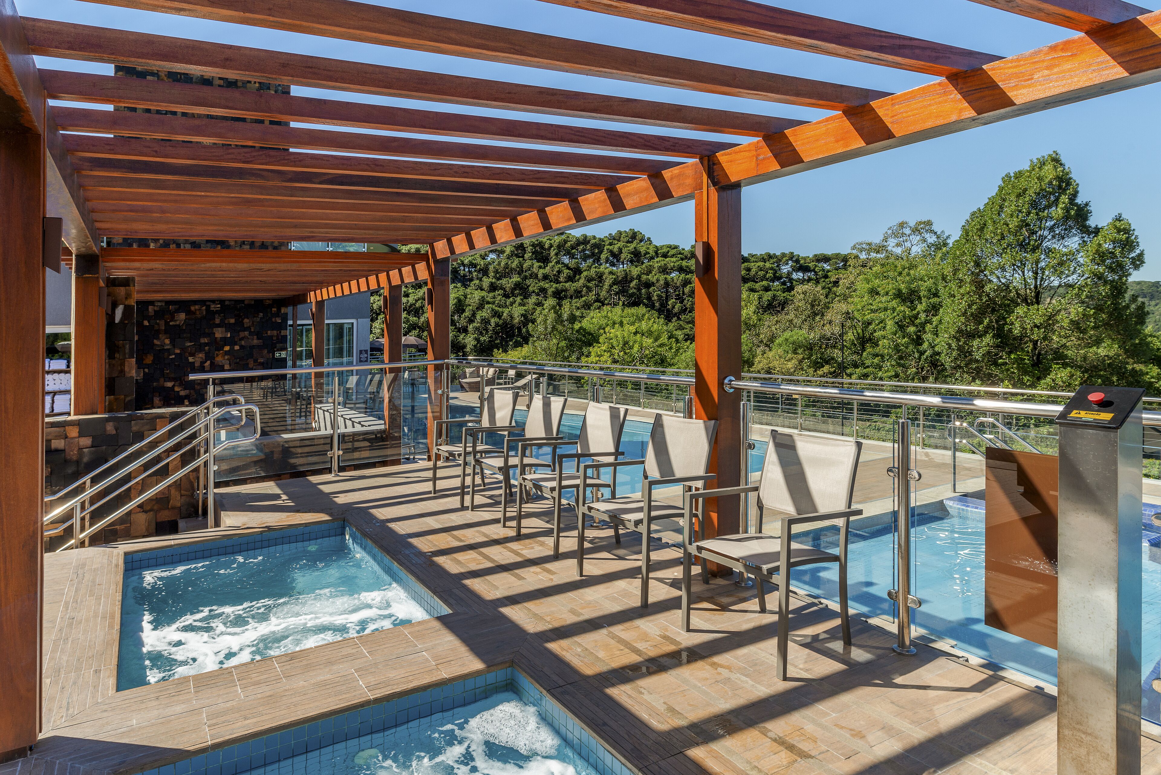 Uma área de piscina com várias banheiras de hidromassagem e espreguiçadeiras, cercada por pérgulas de madeira e um guarda-corpo de vidro.