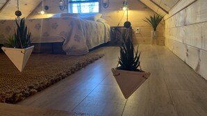Una camera da letto mansardata accogliente, con soffitto spiovente, un letto con copripiumino floreale, due vasi sospesi e una finestra che illumina con luce naturale.
