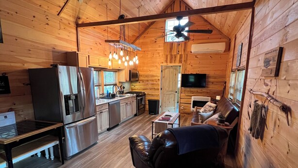 A cozy cabin interior with a wooden interior, a dining table, a sofa, a kitchen area, and a ceiling fan. - LIAHN CABIN AT WINDROCK RIDGE .RIDE TO THE TRAILS IN A MINUTE, RIDE,RELAX,REPEAT (Oliver Springs)