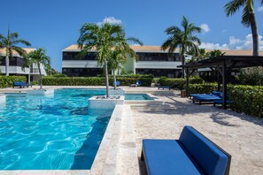 A swimming pool with lounge chairs and palm trees, surrounded by a building with a tiled roof. - Blue Bay Bungalows - The Garden (Sint Michiel)