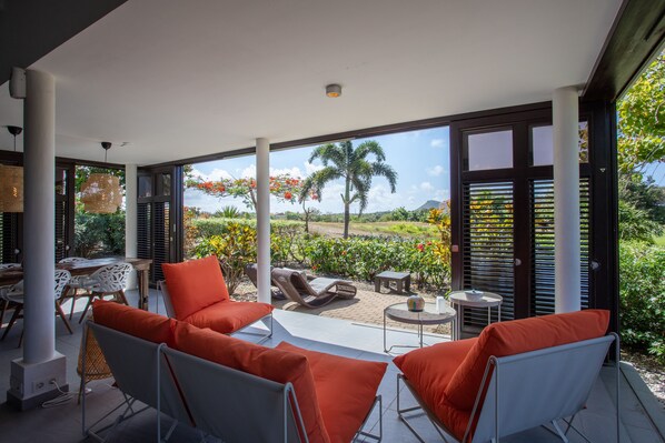 A modern outdoor seating area with orange cushions, a table, and chairs, overlooking a garden with palm trees and flowers. - Blue Bay Bungalows - The Garden (Sint Michiel)