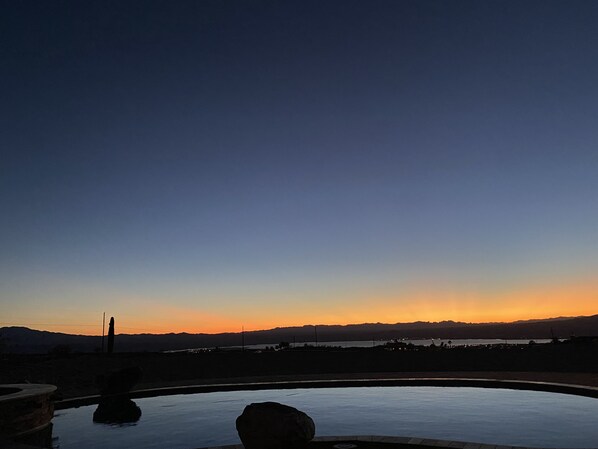 Solnedgang over en swimmingpool med en stensøjle i forgrunden.