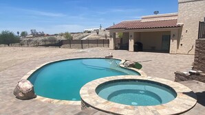 Una piscina con un jacuzzi, rodeada por un patio de ladrillo, y una casa con techo de tejas al fondo.