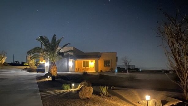 Una casa moderna de noche con un camino de entrada que lleva a la puerta, rodeada de jardines ajardinados y palmeras.