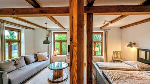 A room with a sofa, a round wooden table, a bed, and a window with a view of greenery. - Ferienwohnung auf Einem Resthof (Quickborn)