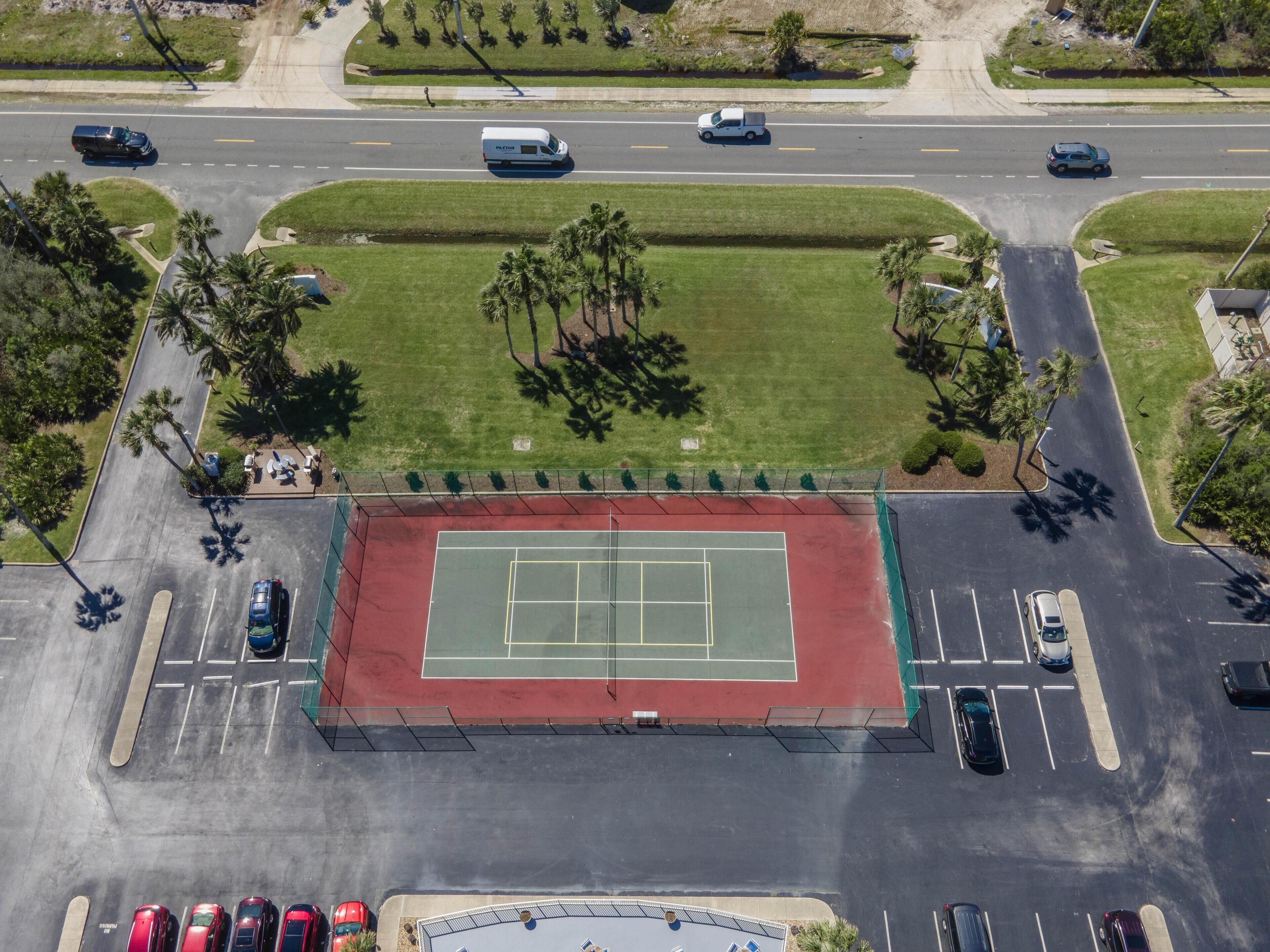 Luftaufnahme eines Tennisplatzes, umgeben von einem Parkplatz und einer Straße mit Fahrzeugen.