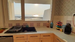 Une cuisine avec une fenêtre donnant sur la plage, un évier, un égouttoir et une bouilloire.