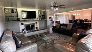A modern living room with a glass coffee table, leather sofas, a flat-screen TV, and a kitchen in the background.