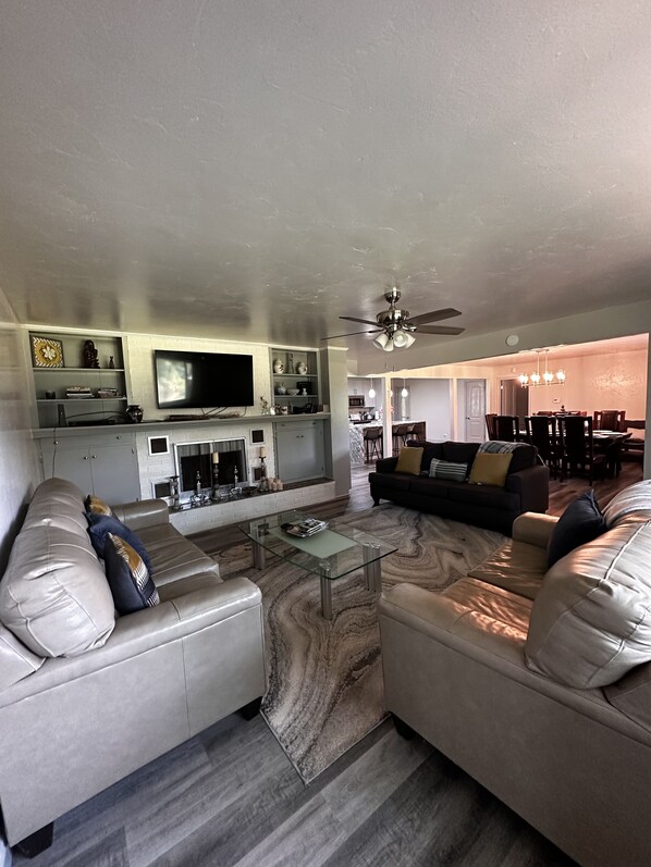 A modern living room with a glass coffee table, leather sofas, a flat-screen TV, and a kitchen in the background. - Amazing lakefront house- DISCOUNTED RATES!!!!!!!!!!!!!!! (Hot Springs)
