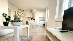 Un salon moderne avec un canapé blanc, une table basse et un téléviseur posé sur un meuble blanc. On aperçoit, au fond, une cuisine avec des meubles blancs et un coin repas avec des chaises blanches.