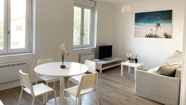 Un salon moderne avec une table de salle à manger ronde, des chaises blanches, un canapé, un téléviseur et un tableau représentant une scène de plage.