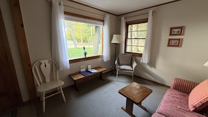Una habitación con un banco de madera, dos sillas, una mesita y un sofá. Hay ventanas con cortinas, una lámpara y cuadros enmarcados en la pared.