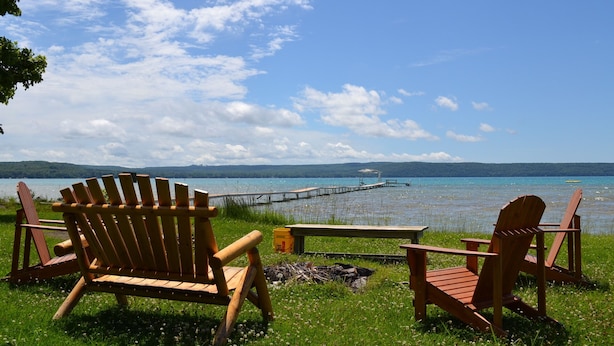Un paisaje junto al lago con sillones de madera tipo Adirondack y una mesa de picnic, un muelle que se extiende sobre el agua, y un cielo despejado.