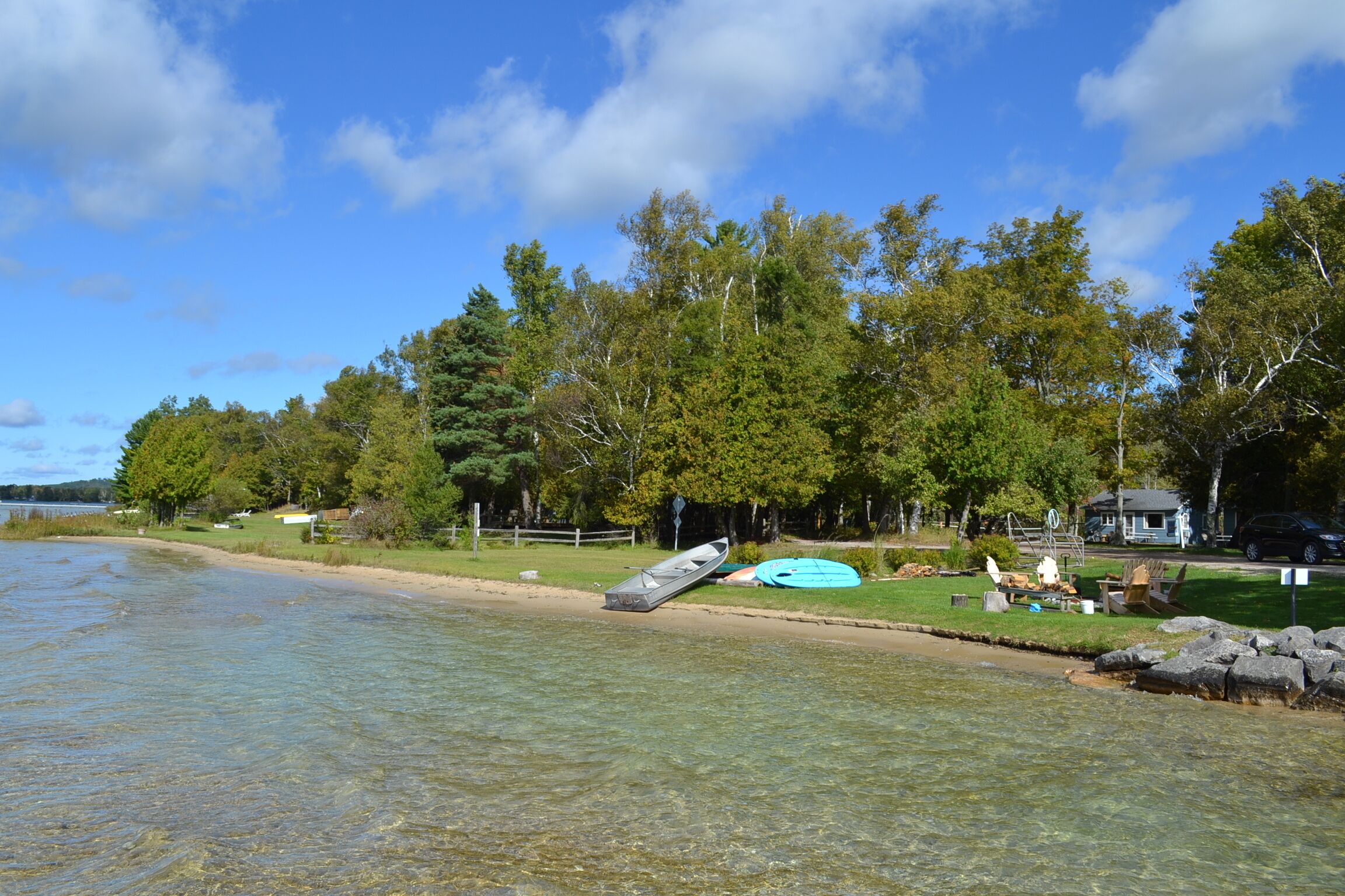 A lakeside property with clear water, a sandy beach, and a row of trees.
