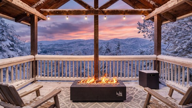 Une terrasse en bois avec un foyer, des sièges et une vue sur les montagnes enneigées au coucher du soleil.