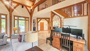 A spacious living area with a high ceiling, wooden beams, a sofa, a desk with a computer, and a view of the outdoors.