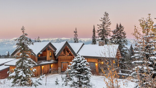 A log cabin with a symmetrical facade, multiple chimneys, and a snow-covered landscape with mountains in the background.