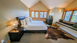 A bedroom with a large bed, a wooden headboard, a nightstand with a lamp, a dresser, a television, a window seat, and a fur rug. - Creekside Chalet: 7-bedroom luxury mountain home (Golden)