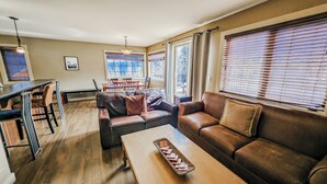 Een ruime woonkamer met een bruine leren bank, een houten salontafel en een eethoek met een tafel en stoelen.