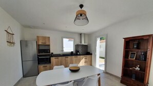 A modern kitchen with a dining area, featuring a white table, chairs, and a wooden cabinet. - L’embrun           . (Dolus-d'Oléron)