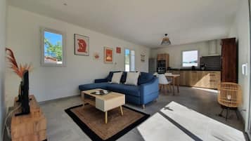 Un salon moderne avec un canapé, une table basse et un coin repas avec une chaise. On aperçoit une cuisine à l’arrière-plan, avec des placards et une fenêtre.