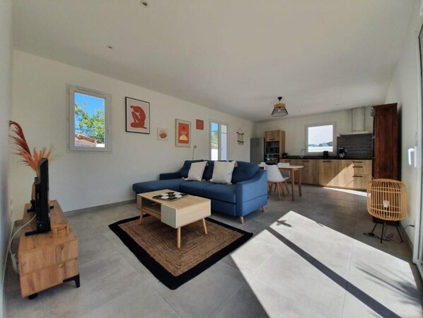 A modern living room with a sofa, coffee table, and a dining area with a chair. There is a kitchen in the background with cabinets and a window. - L’embrun . (Dolus-d'Oléron)