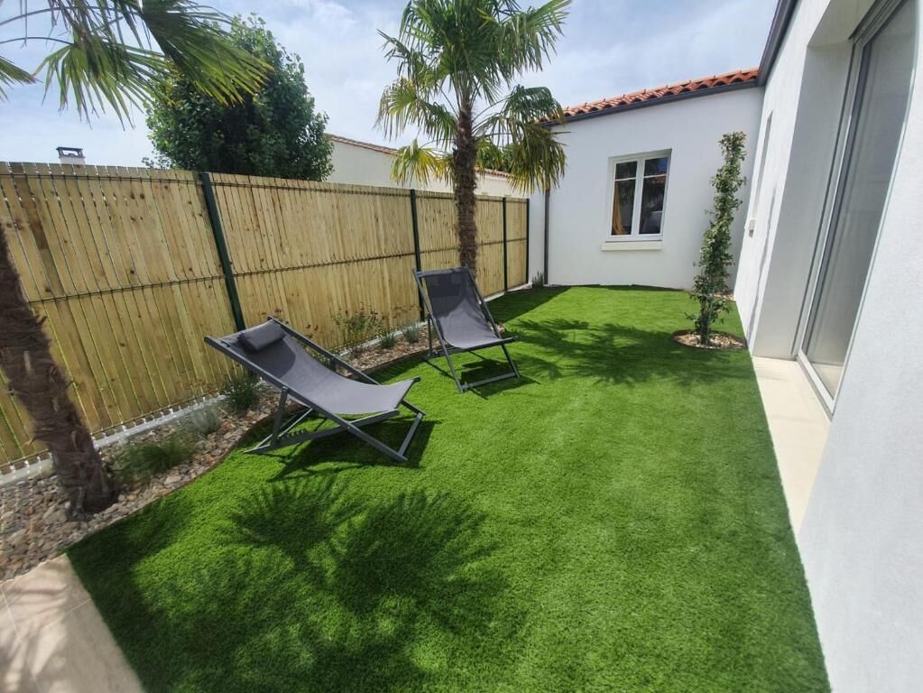A backyard with artificial grass, two lounge chairs, a wooden fence, and a small palm tree.
