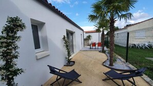 A courtyard with two lounge chairs, a palm tree, and a white building with a tiled roof. - L’embrun           . (Dolus-d'Oléron)
