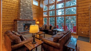 L’intérieur d’une cabane en rondins est agrémenté d’une cheminée en pierre, de canapés en cuir et de grandes fenêtres qui offrent une vue sur les arbres.