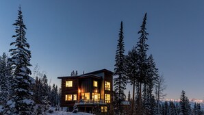Una cabaña moderna de madera con múltiples ventanas, rodeada de árboles nevados y un paisaje montañoso.