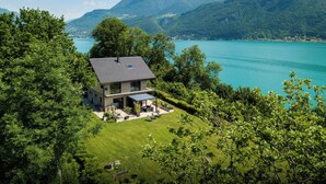 Il s’agit d’une maison moderne à deux étages, dotée d’un toit plat, de grandes fenêtres et d’un patio aménagé avec du mobilier d’extérieur. Elle est entourée d’une végétation luxuriante et offre une vue sur un lac turquoise, avec des montagnes en toile de fond.