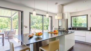Une cuisine moderne dotée d’un grand îlot central, d’appareils électroménagers en acier inoxydable et de grandes fenêtres offrant une vue sur la verdure.