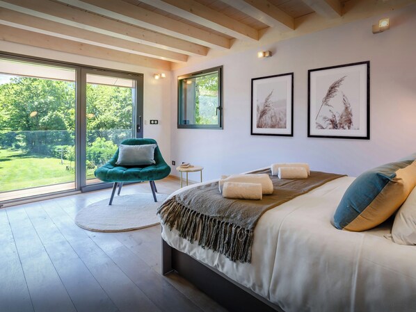 Ein modernes Schlafzimmer mit einem großen Bett, einem grünen Sessel und Blick auf einen Garten durch eine Schiebetür.