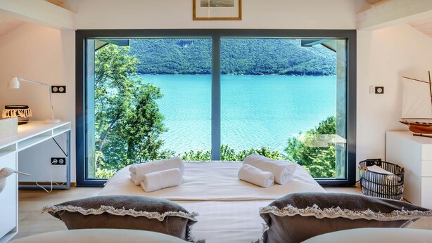 Une chambre avec une grande fenêtre donnant sur un lac, un lit avec des oreillers, un bureau avec une lampe et une petite maquette de bateau posée sur une étagère.
