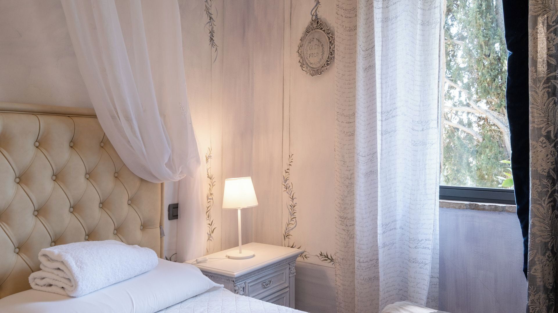 A bedroom with a tufted headboard, a bedside table with a lamp, and a window with curtains.