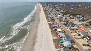 An aerial view of a coastal residential area with houses along the beach, a road, and a clear sky. - Oceanfront family fun home and dog friendly! Coastal Coop (Oak Island)