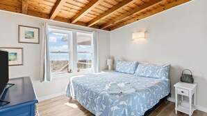 A bedroom with a bed, a nightstand, a lamp, and a window with curtains. - Oceanfront family fun home and dog friendly! Coastal Coop (Oak Island)