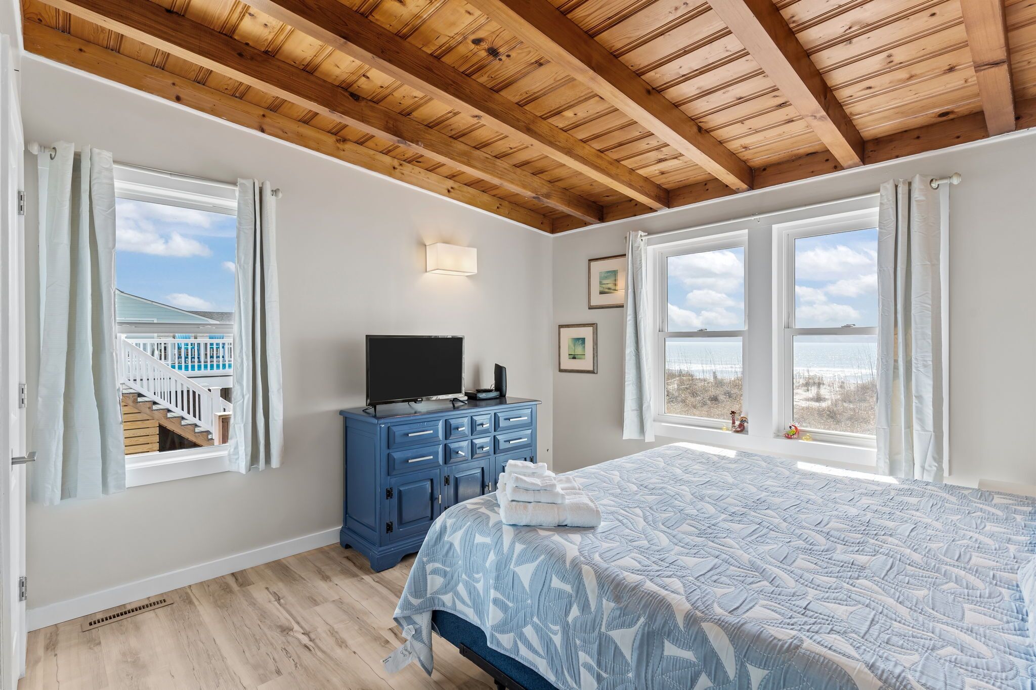 Ein Schlafzimmer mit Holzboden, einem Bett mit gemustertem Überwurf, einer blauen Kommode, einem Fernseher und großen Fenstern mit Blick auf ein Gebäude und den Himmel.